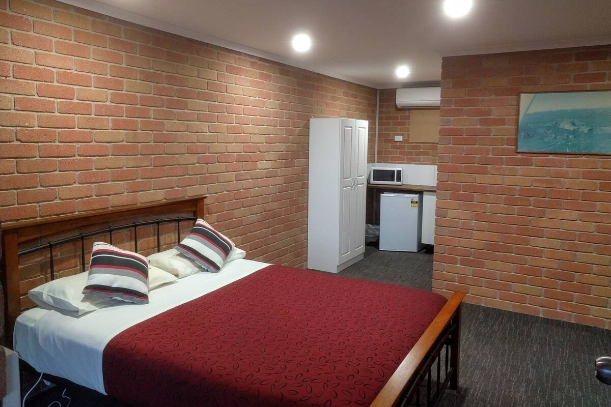 A room with a brick wall, a bed with pillows, a red blanket, a white wardrobe, a small kitchenette with a microwave, and a painting on the wall.