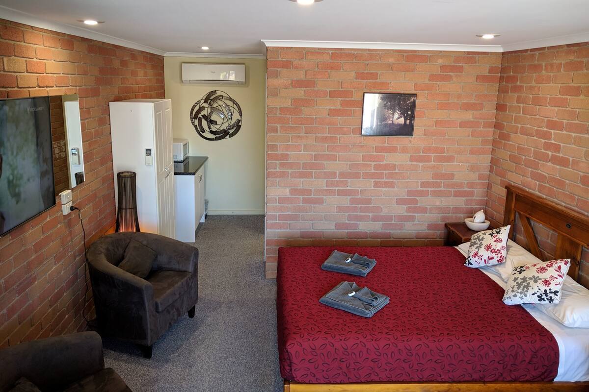 A bedroom with a brick wall, a bed with a red bedspread, a wooden headboard, and a small bedside table with a lamp.