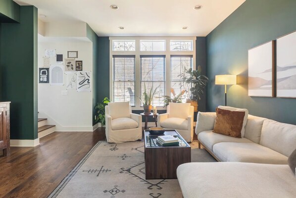 A living room with a sofa, two armchairs, a coffee table, and a rug. There are windows with blinds, a lamp, and wall art.
