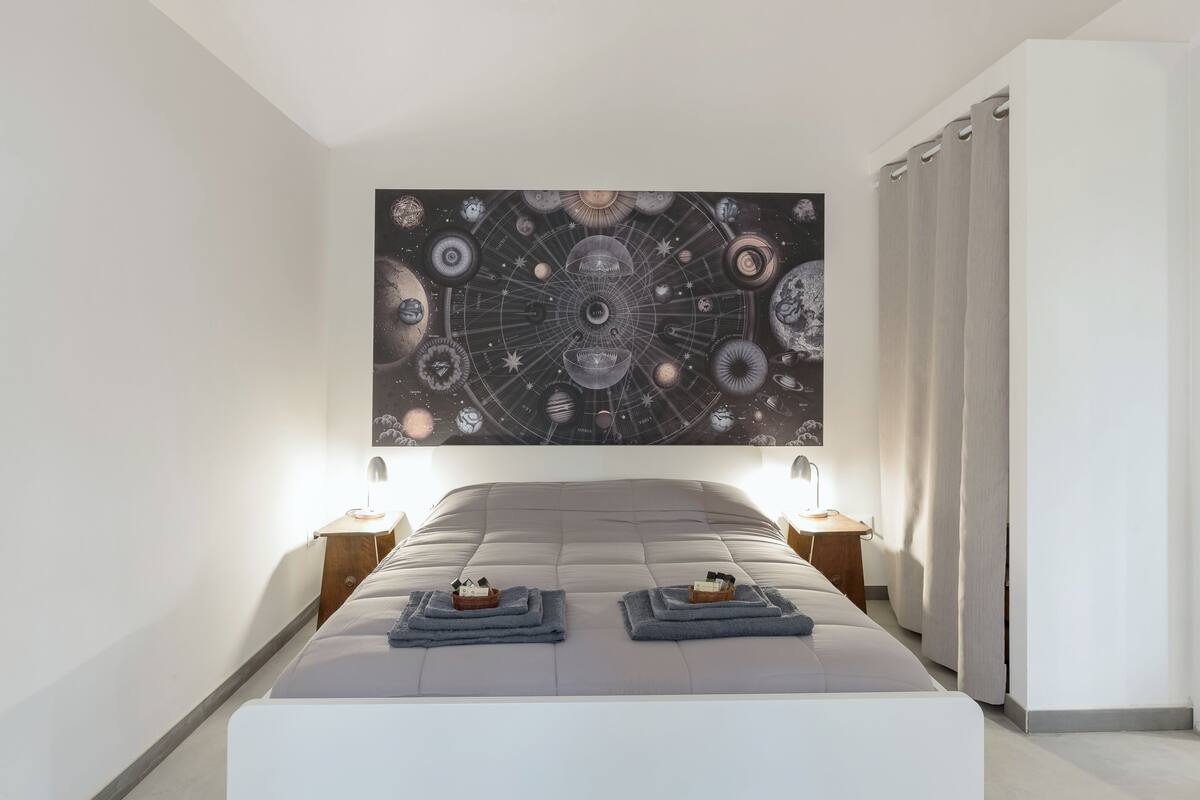 A bedroom with a large wall tapestry of celestial bodies, a bed with two nightstands, and a door to another room.