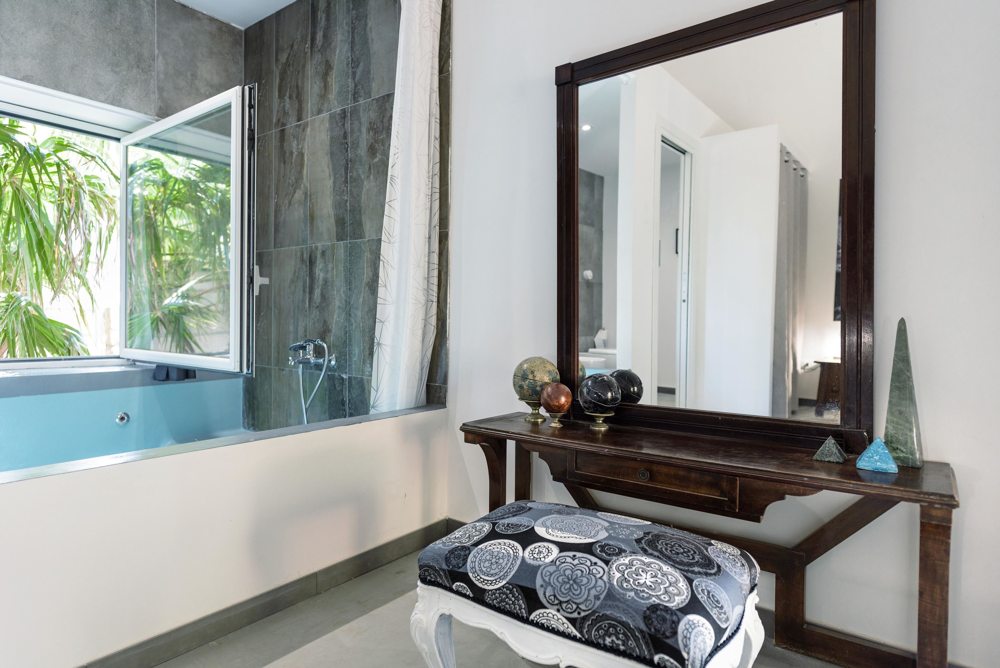 A bathroom with a bathtub, a large mirror, a vanity with decorative items, and a window with a view of greenery.