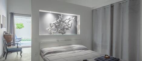 A modern bedroom with a large bed, a chair, a window with curtains, and a wall-mounted abstract art piece.
