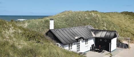 Ein weißes Gebäude mit schwarzem Dach, umgeben von Gras und Dünen, mit Blick auf das Meer.