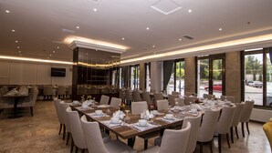 A spacious dining area with multiple tables set for service, large windows offering a view of the street, and a modern ceiling design with recessed lighting. - An Grand Hotel (Ankara)