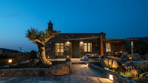 Ein Steinhaus mit einem großen Baum davor, eine Terrasse mit Gartenmöbeln und Blick auf das Meer.