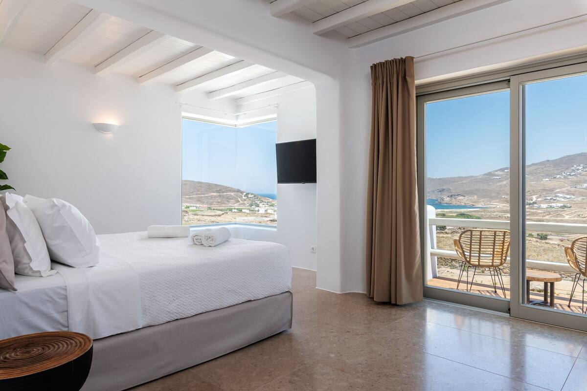 A bedroom with a bed, a TV, a balcony with chairs, and a view of the landscape.