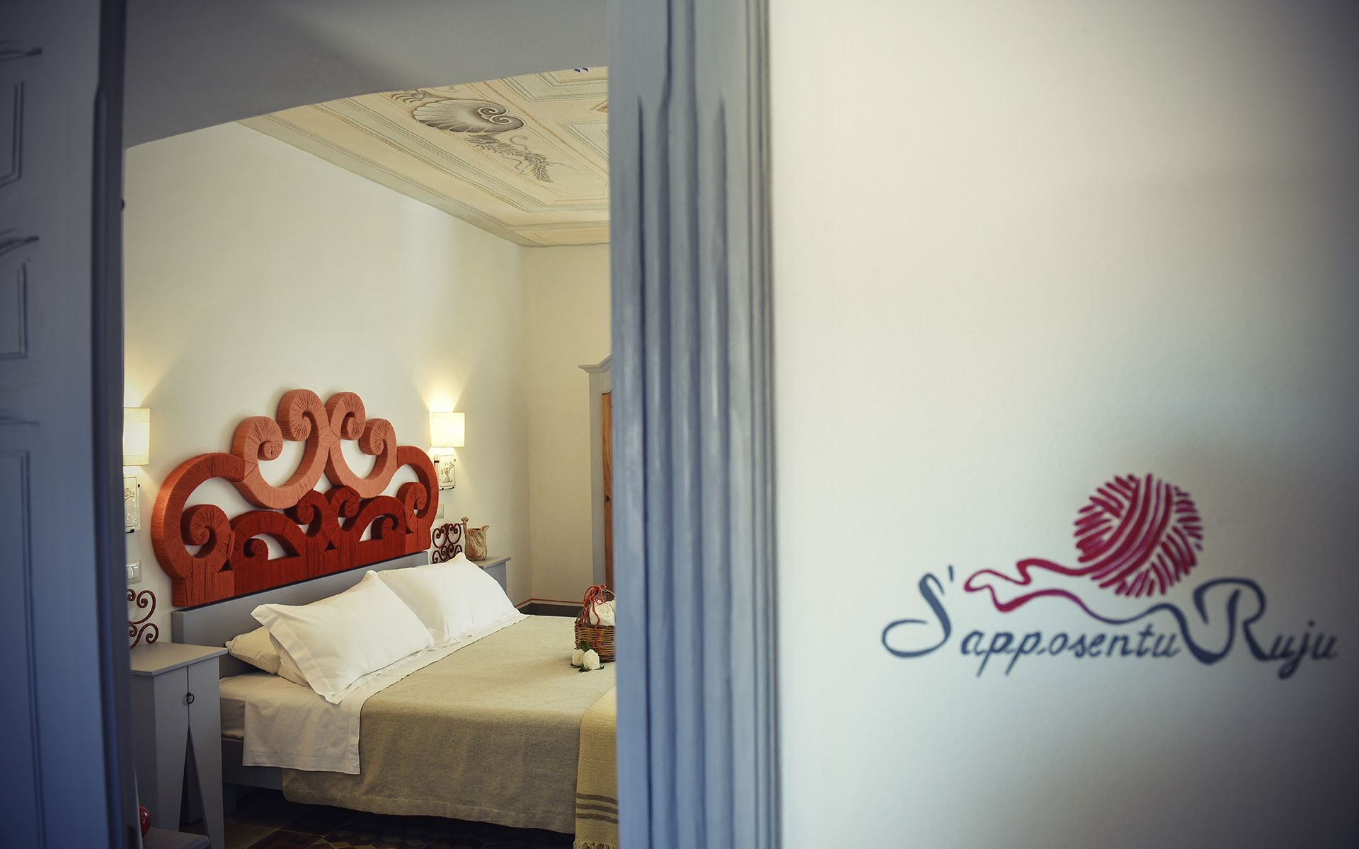 Un dormitorio con cama, cabecera decorativa, mesita de noche y una pared con un logo.