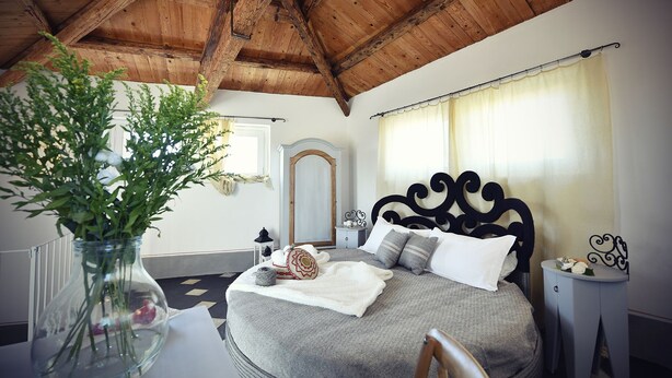 A bedroom with a wooden ceiling, a bed with a black headboard, a window with curtains, and a vase with green plants on a table. - Bisos Ospitalità diffusa eco-sostenibile (Paulilatino)