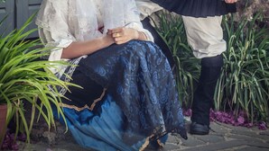 A person in traditional attire sitting on the ground with a stone wall and a door in the background. - Bisos Ospitalità diffusa eco-sostenibile (Paulilatino)