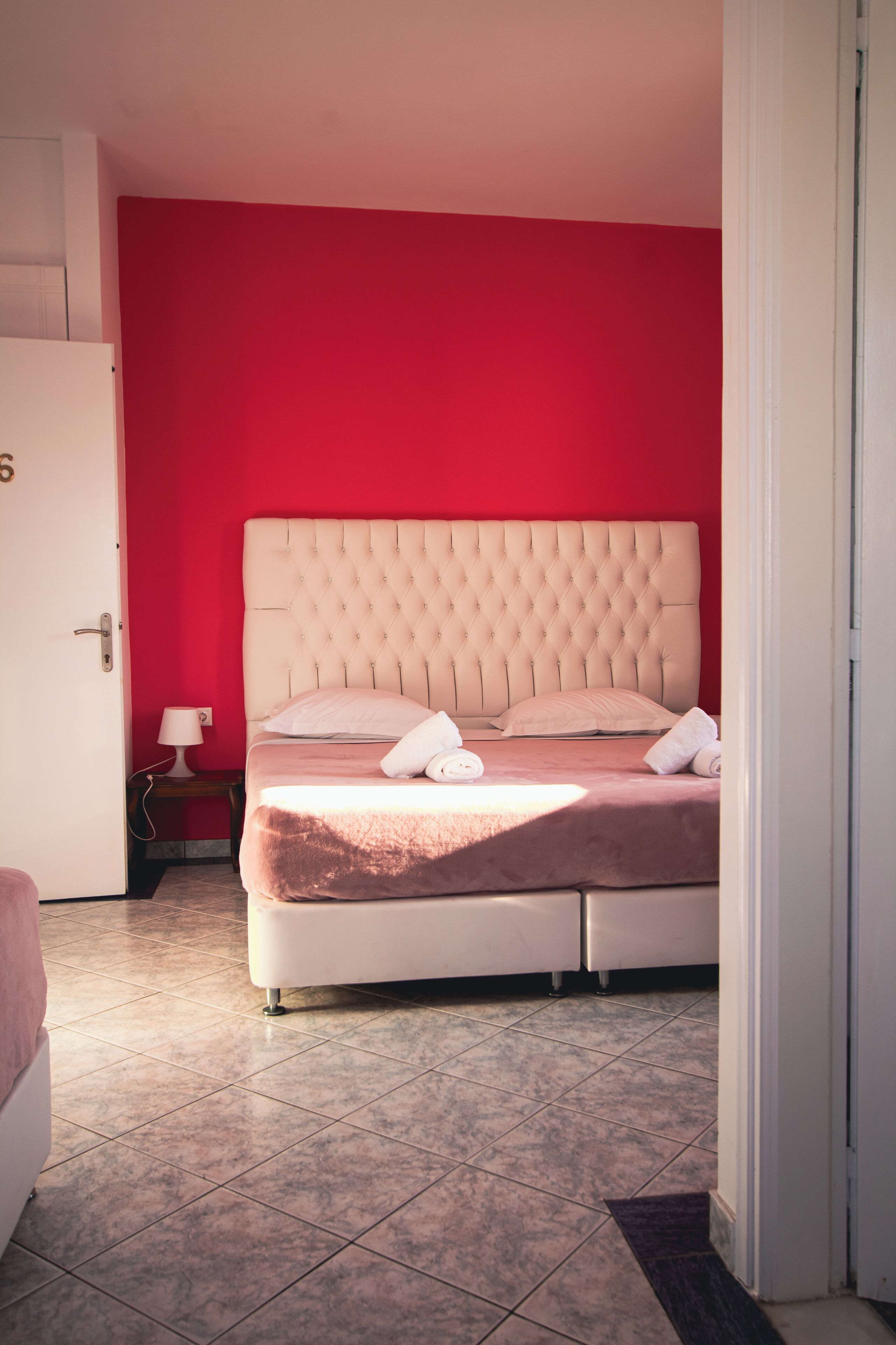 Habitación de hotel con pared roja, cabecera capitonada, cama con ropa blanca y mesita de noche con lámpara.