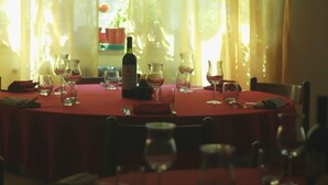 Une table à manger dressée avec des verres à vin, une bouteille et un pichet, agrémentée de rideaux légers et offrant une vue sur la verdure à l’extérieur.