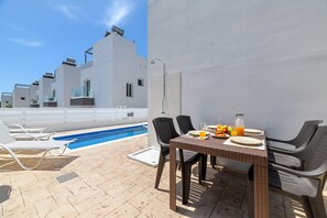 Un espace piscine sur le toit, avec des chaises longues, une table à manger avec des assiettes et une bouteille, et un ciel bleu dégagé.