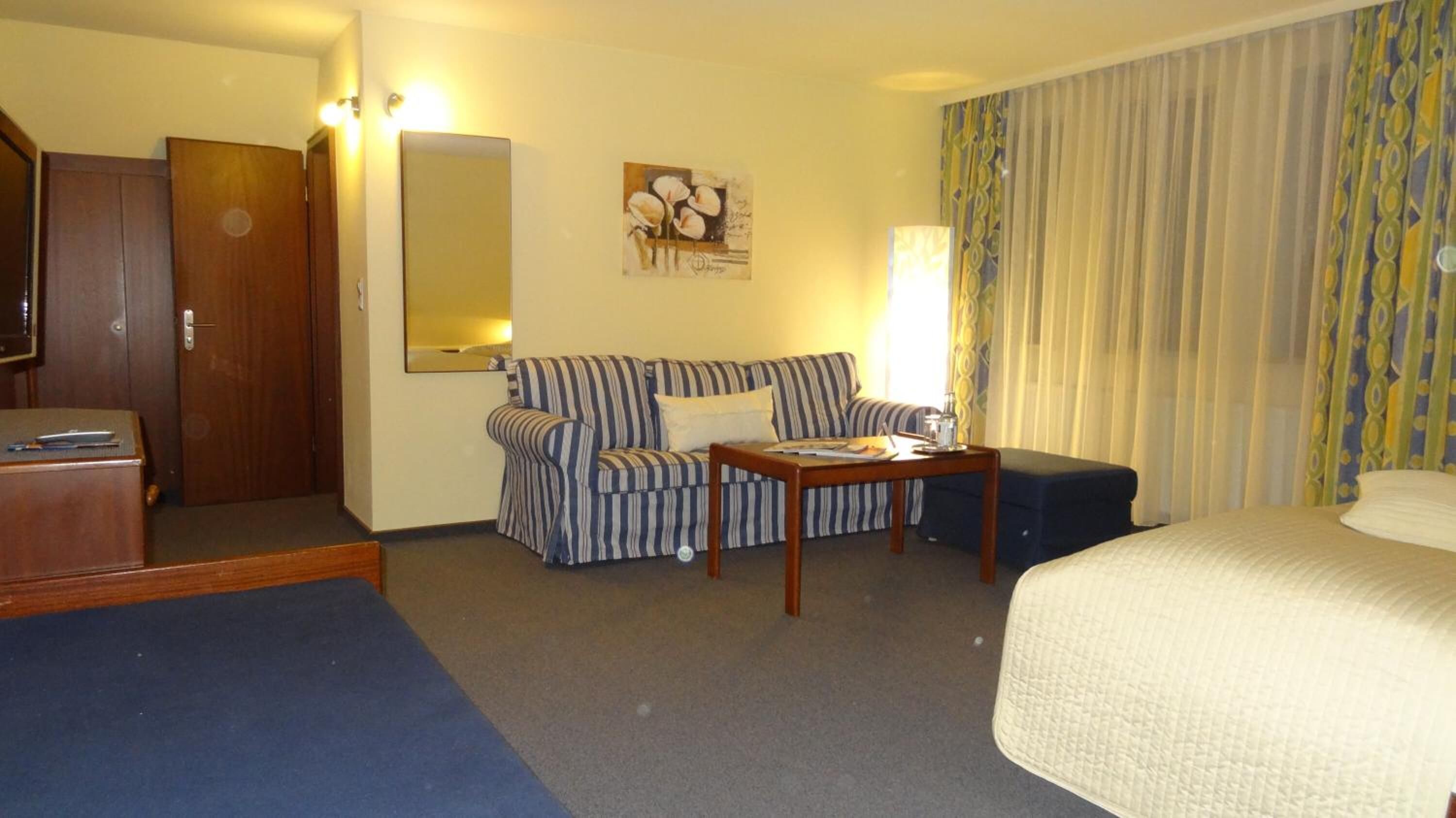 A hotel room with a bed, a striped sofa, a small table with a lamp, a wooden desk, and a large mirror.