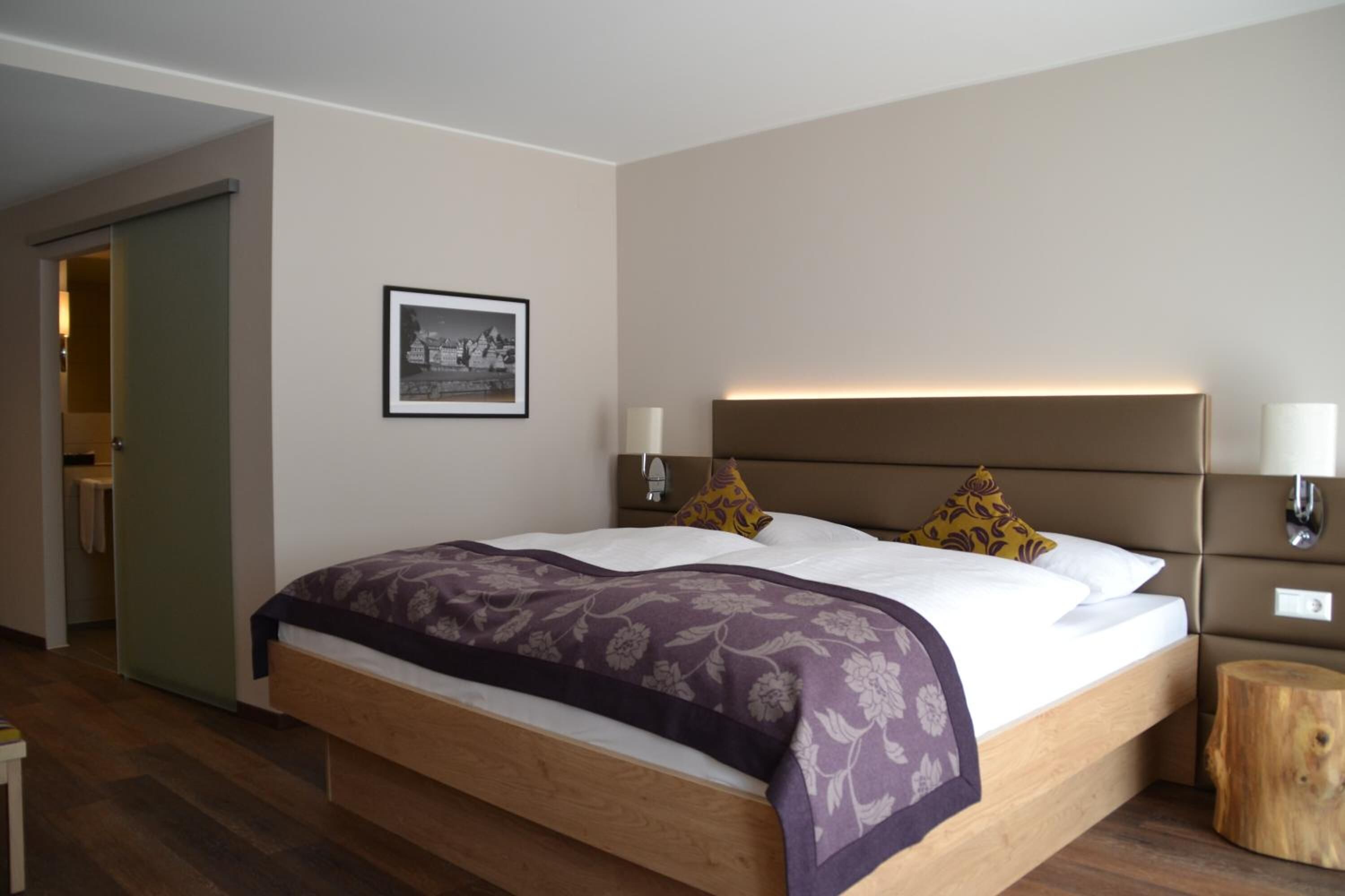 Een moderne hotelkamer met een houten bed, een hoofdbord met ingebouwde verlichting, een nachtkastje met een lamp en een ingelijst schilderij aan de muur.