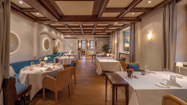 A restaurant with a long, narrow layout featuring high ceilings with wooden beams, white tablecloths, and wicker chairs.