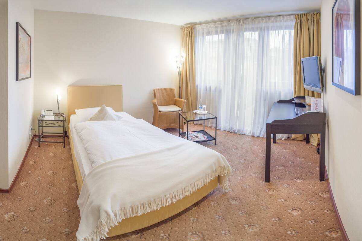 Ein Hotelzimmer mit Bett, Sessel, Schreibtisch, Fernseher und einem fensterbepflanzten Fenster.