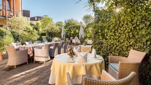 An outdoor dining area with wicker furniture and round tables, surrounded by greenery and buildings.