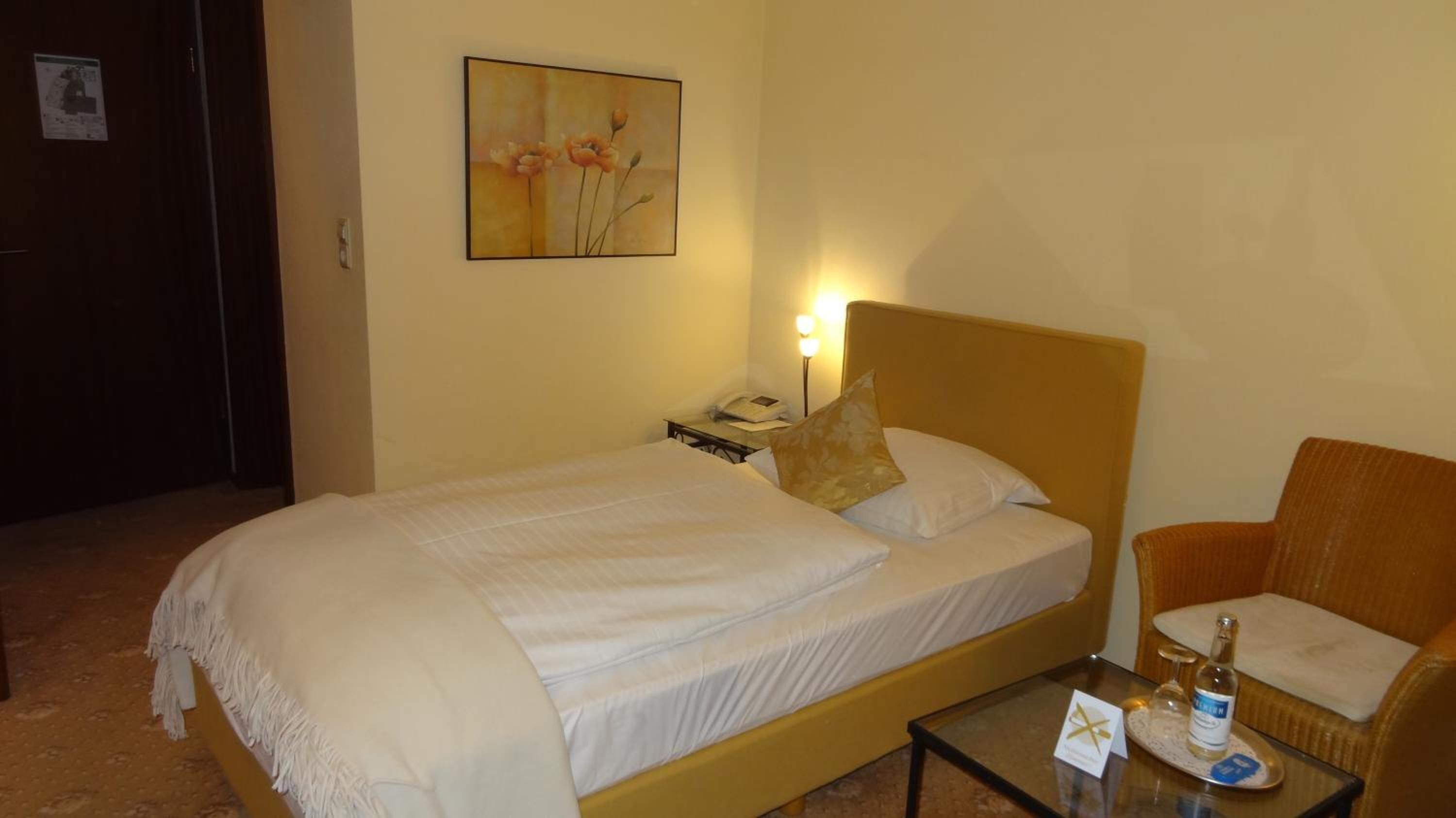 Habitación de hotel con una cama, una mesita de noche, una lámpara, una silla y una pequeña mesa con una botella y una tarjeta.