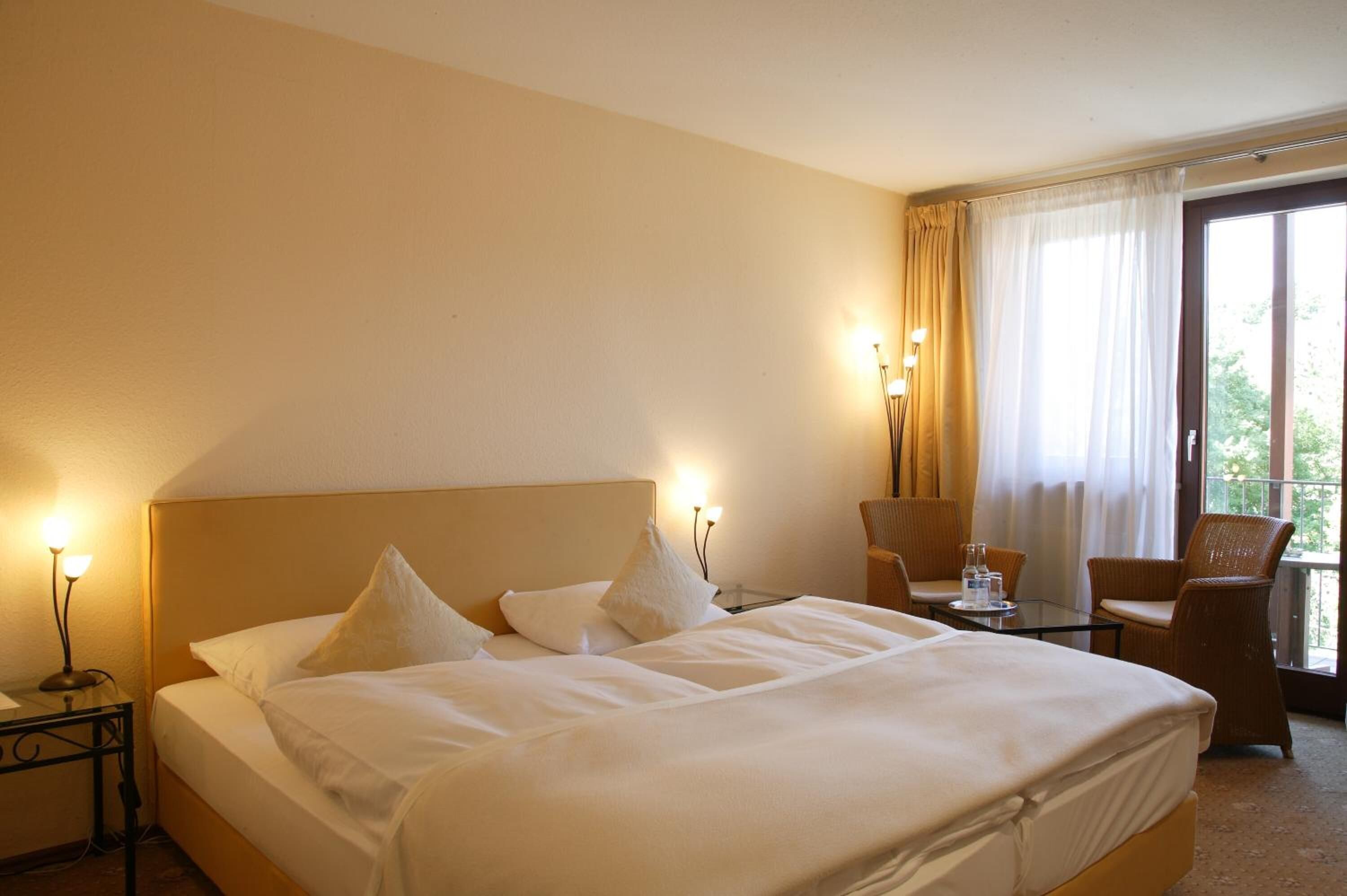 A hotel room with a large bed, two chairs, a small table, and a window with curtains.