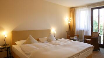 A hotel room with a large bed, two chairs, a small table, and a window with curtains.