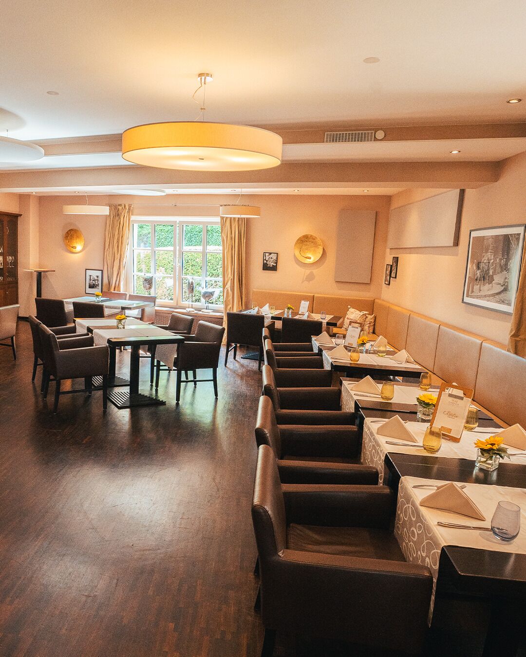 A restaurant with a long counter, multiple tables, and chairs. There are windows with curtains, a chandelier, and framed pictures on the walls.