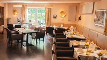 A restaurant with a long counter, multiple tables, and chairs. There are windows with curtains, a chandelier, and framed pictures on the walls.
