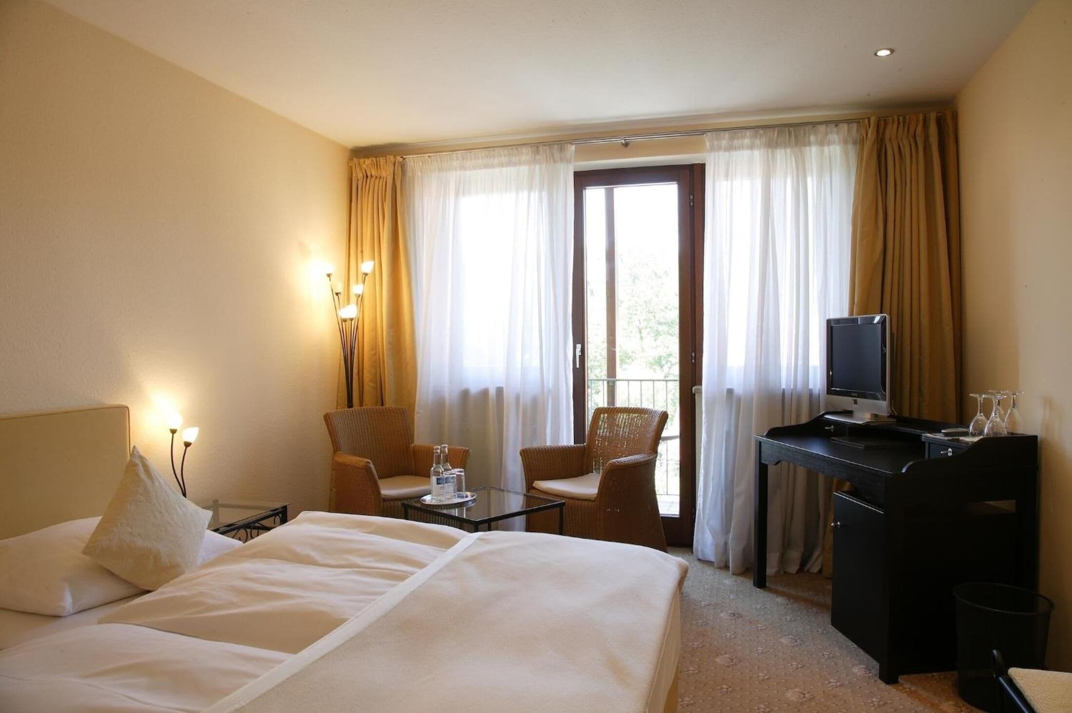 A hotel room with a bed, a desk with a computer, two chairs, a small table with a bottle, and a window with curtains.