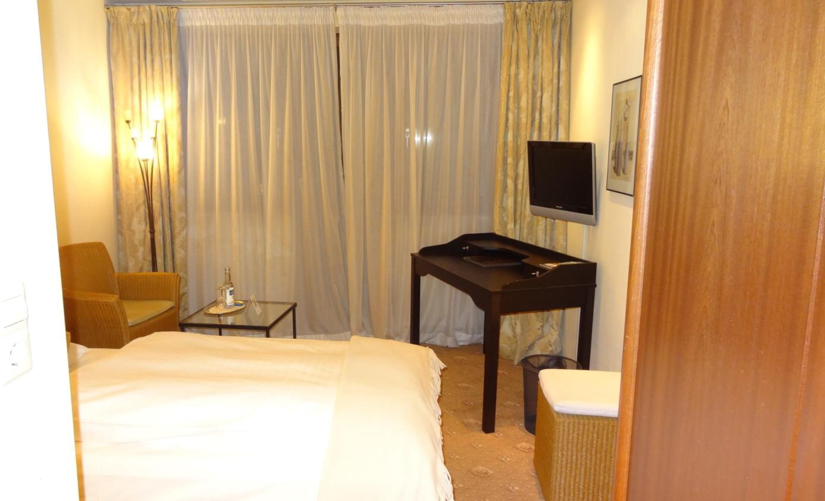 Une chambre d’hôtel avec un lit, une petite table sur laquelle se trouve une bouteille, une chaise, un téléviseur et des rideaux.