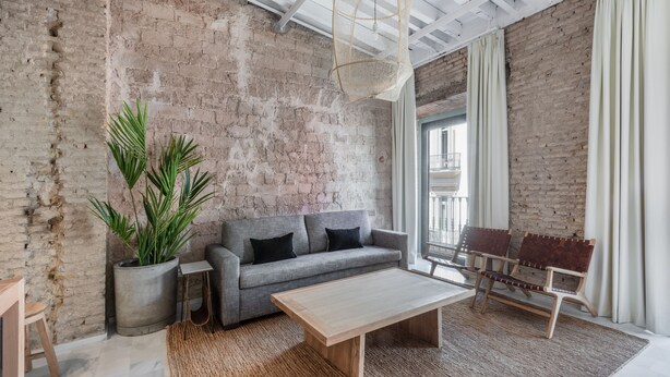 Uma sala de estar com um sofá cinza, uma mesa de centro de madeira, um tapete tecido, uma planta em vaso e um banco com assento de fibra natural.