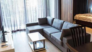 Una habitación de hotel moderna con un sofá gris, una mesita blanca pequeña y una cama con colcha color crema.