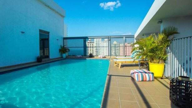 Una piscina en la azotea con agua azul cristalina, rodeada por un edificio blanco y una barandilla de cristal.