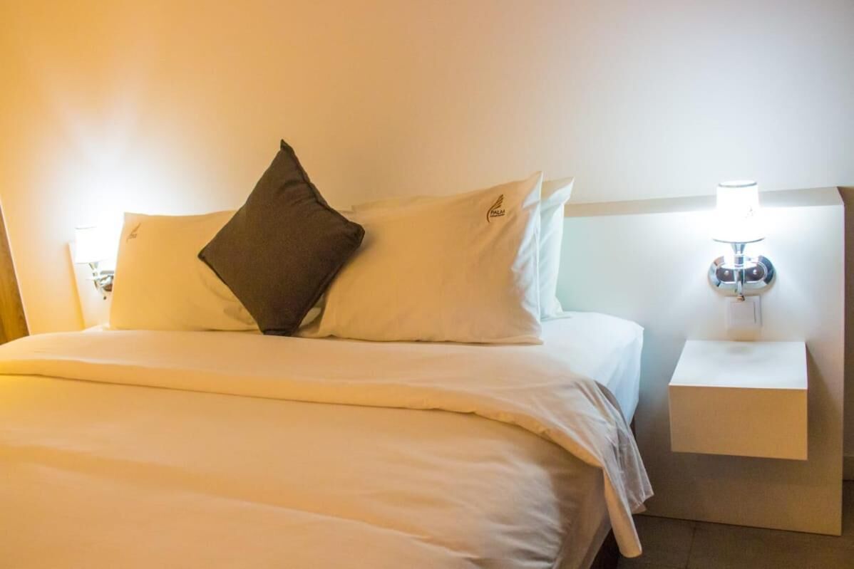 A neatly made bed with white bedding and a dark pillow, a bedside table with a lamp, and a wall-mounted light fixture.