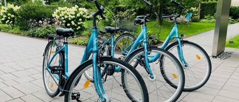 A row of turquoise bicycles parked on a paved surface with a garden and trees in the background.
