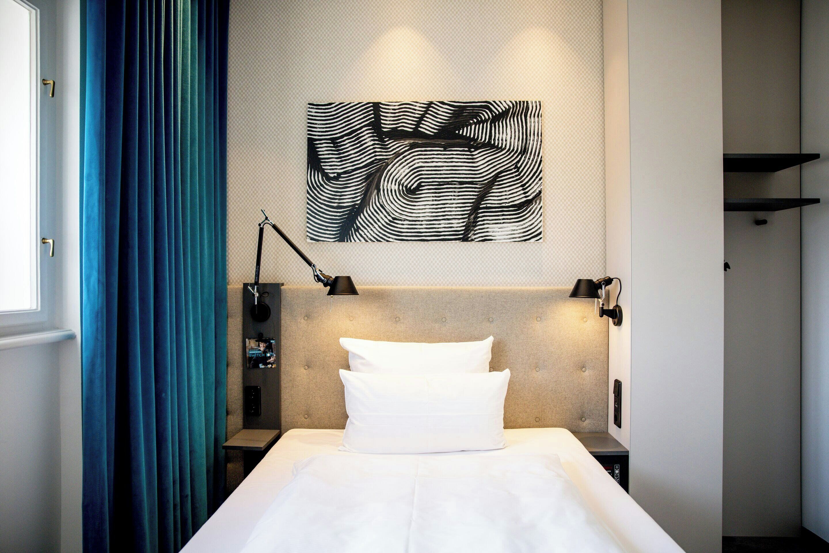 A hotel room with a bed, a headboard, a wall-mounted artwork, a lamp, and a window with curtains.