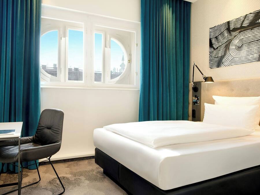 A hotel room with a bed, a desk, a chair, a window with curtains, and a wall-mounted lamp.