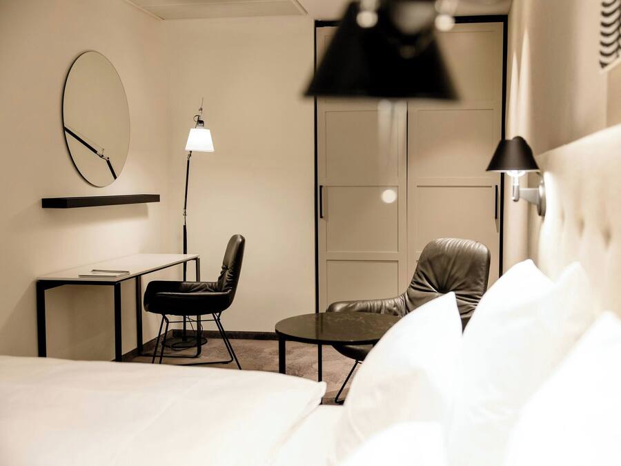 A hotel room with a bed, a desk, two chairs, a round table, a lamp, and a wardrobe.