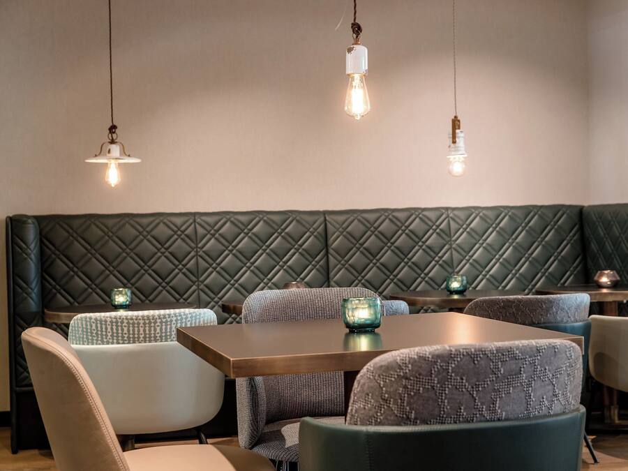 A restaurant interior with a green quilted bench, patterned chairs, and hanging pendant lights.