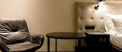 Habitación de hotel con una silla de cuero, una mesita redonda pequeña, una cama con ropa blanca, un cabecero con una lámpara y un panel de control.
