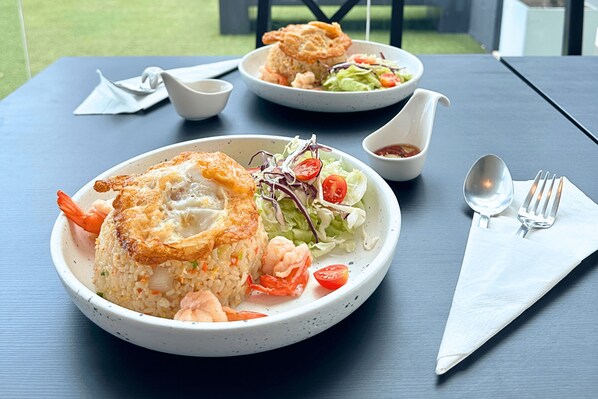 A table with a plate of fried rice topped with a fried egg, shrimp, and vegetables, accompanied by a side salad and a bowl of sauce.