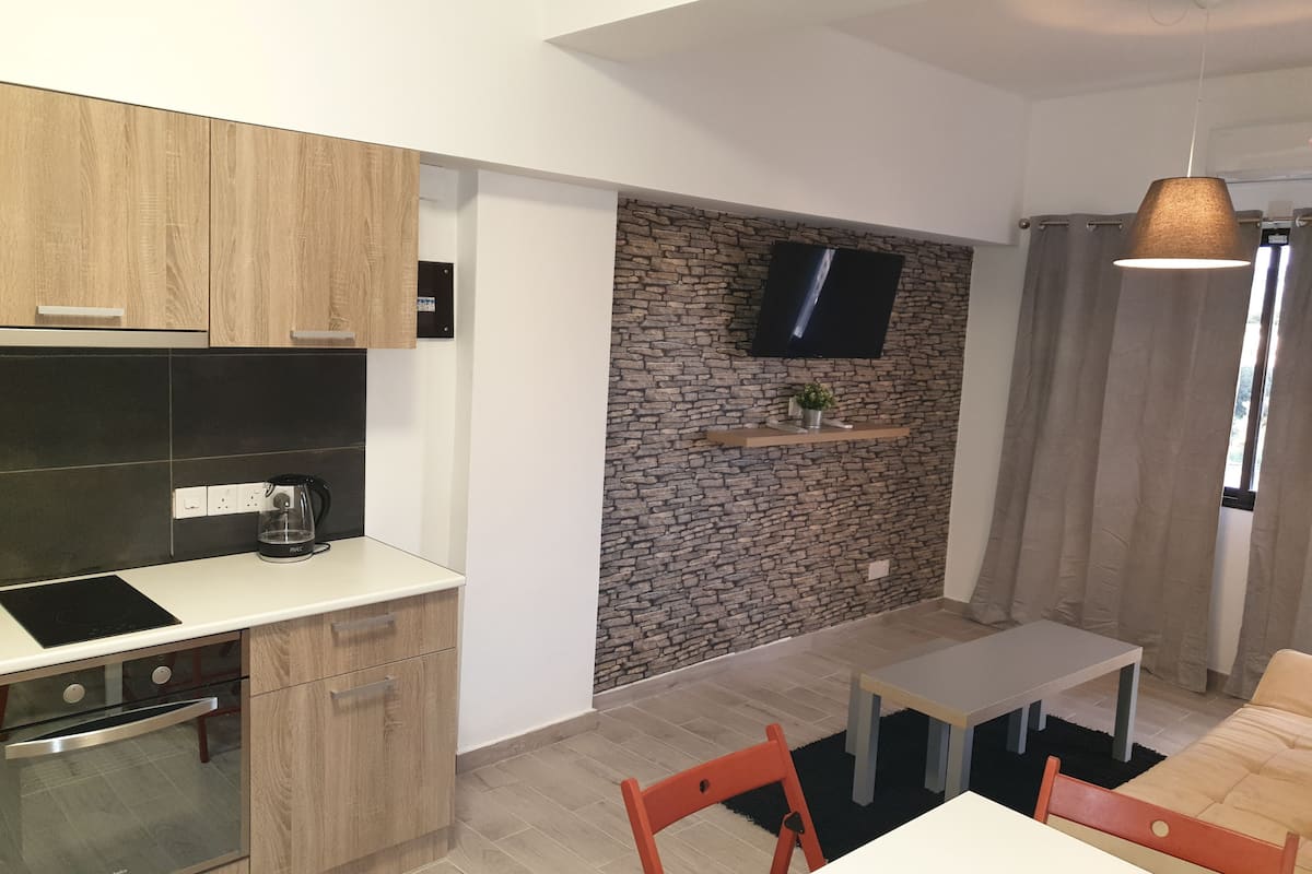 A modern kitchen with wooden cabinets, a built-in oven, and a coffee maker. There is a stone wall with a TV mounted above it and a small shelf with a plant. A dining area with a table and chairs is visible.