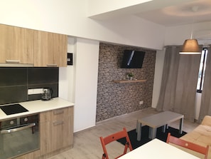 A modern kitchen with a built-in oven and a coffee maker, a stone wall with a TV mounted above, and a dining area with a table and chairs. - Mylos Court Apartments (Limassol)