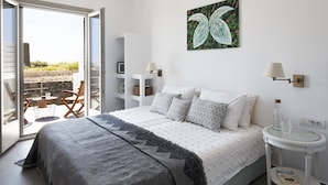 A bedroom with a bed, a side table, a chair, and a view of the outdoors. - Alchemy Luxury Villa (Santorini)