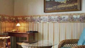 Ein Zimmer mit gestreifter Tapete, einem Bild an der Wand, einer Lampe und einem Korbstuhl.