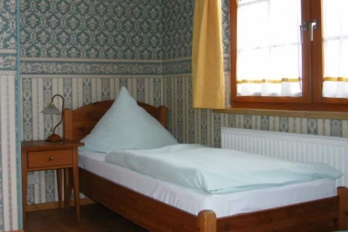 Una cama individual con estructura de madera y ropa de cama blanca en una habitación con papel tapiz estampado y una ventana con cortinas amarillas.