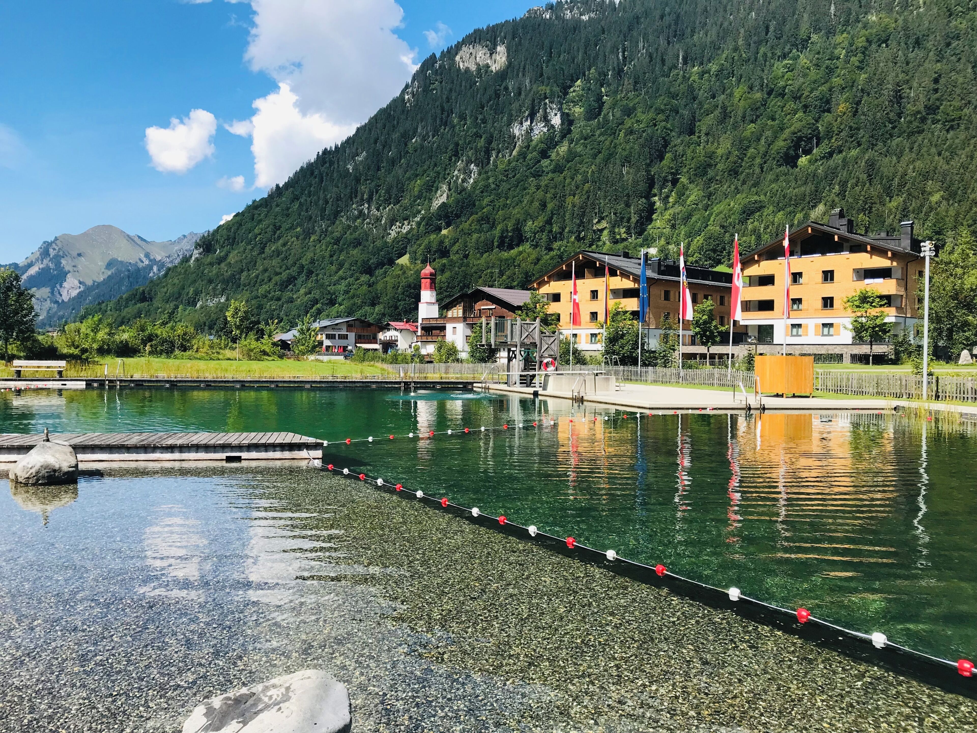 A lakeside hotel with a dock, buildings with balconies, and a mountain backdrop.