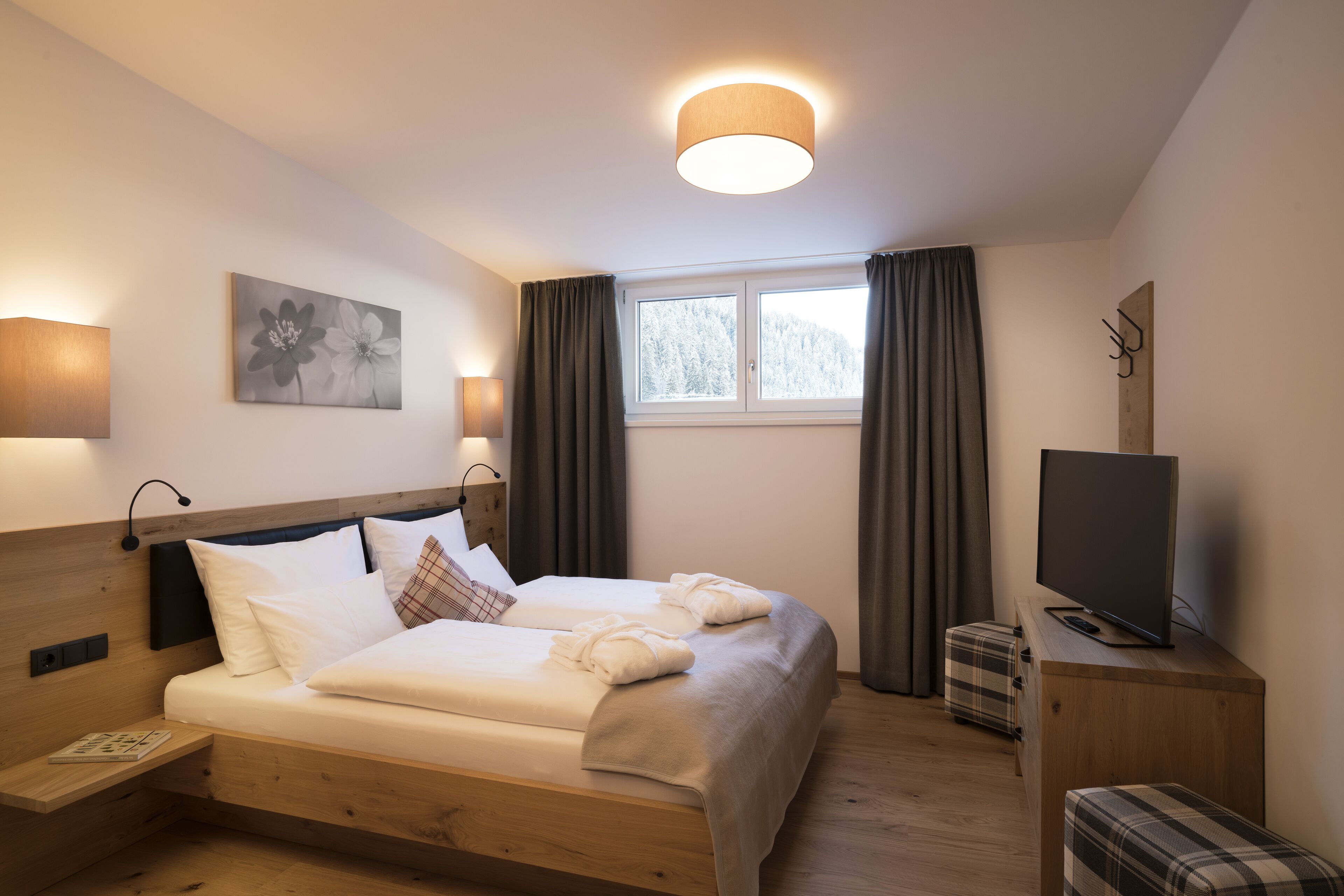Ein Hotelzimmer mit einem hölzernen Bett, einem Flachbildfernseher, einem Nachttisch und einem Fenster mit Vorhängen.