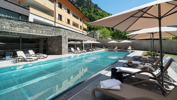 A swimming pool with lounge chairs and umbrellas, a stone facade building, and a mountain in the background. - Arlberg Resort Klösterle (Klösterle am Arlberg)
