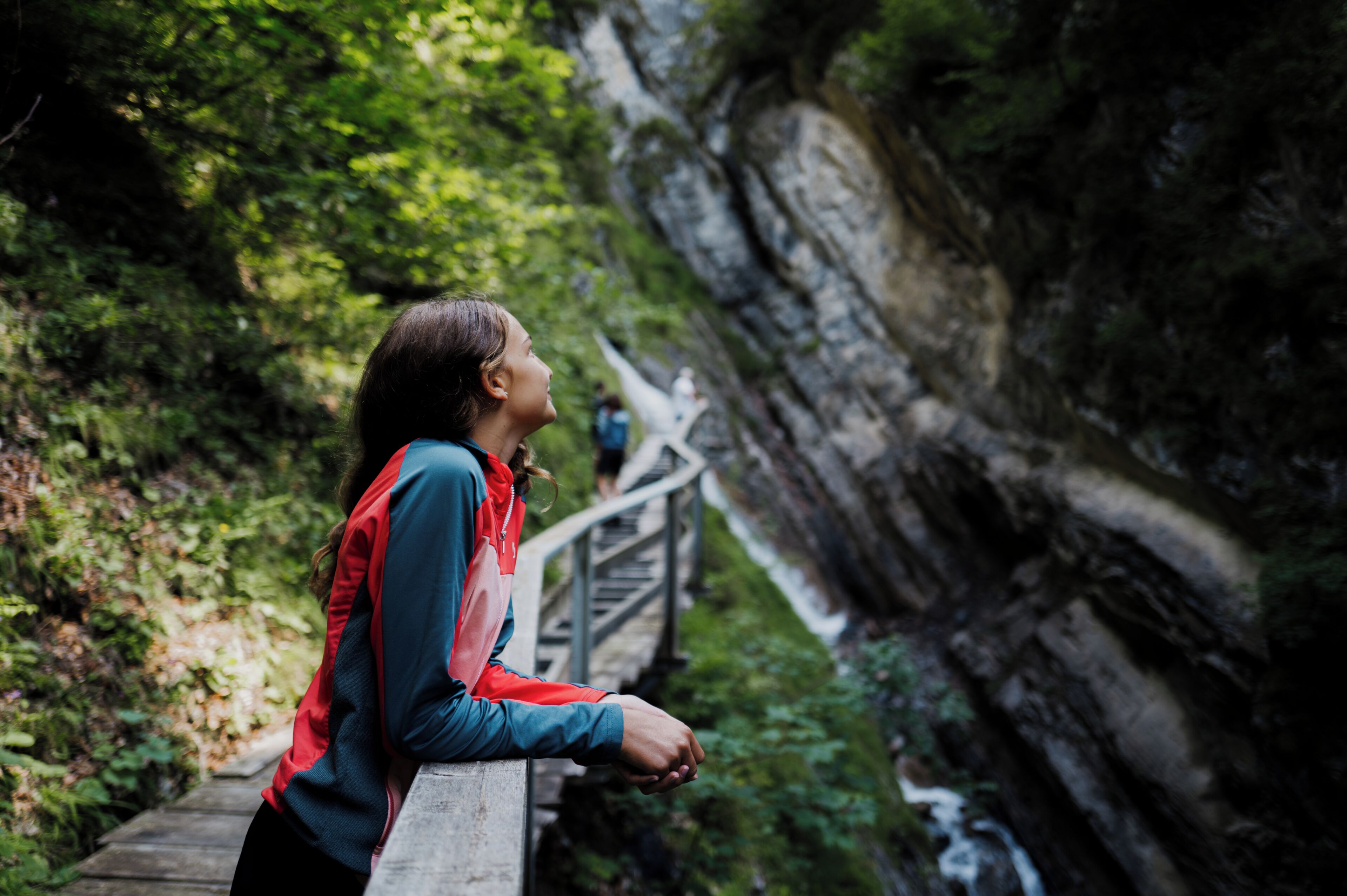 Eine Person steht auf einer Holzbrücke in einem bewaldeten Gebiet mit einer felsigen Klippe.