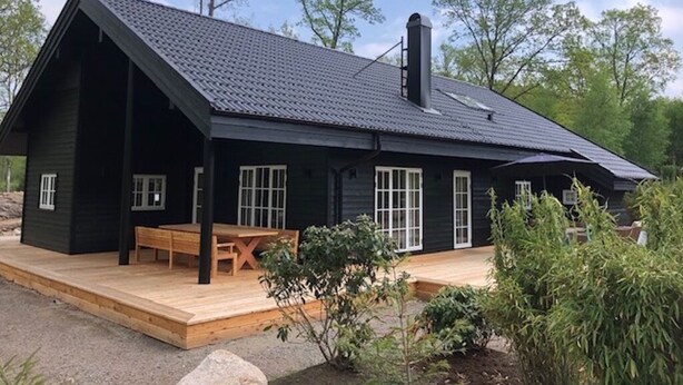A black cabin with a sloped roof, a wooden deck, and a chimney. - Stylish Lake house, outdoor hottub Lake view Close to nature. Pet friendly. (Våxtorp)