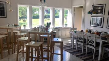 A bright dining area with wooden furniture, large windows, and a view of greenery outside.